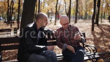 父亲和成年儿子坐在秋天公园的长凳上聊天。 与年迈的父亲在一个